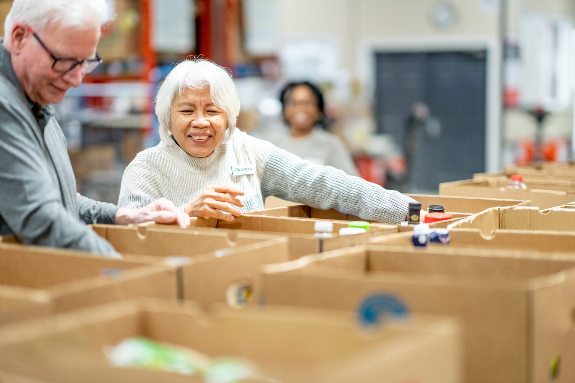 A senior couple are seen volunteering their time at a local food bank as they work together to pack boxes of donations.  They are both dressed casually and smiling as they enjoy giving back to their community.